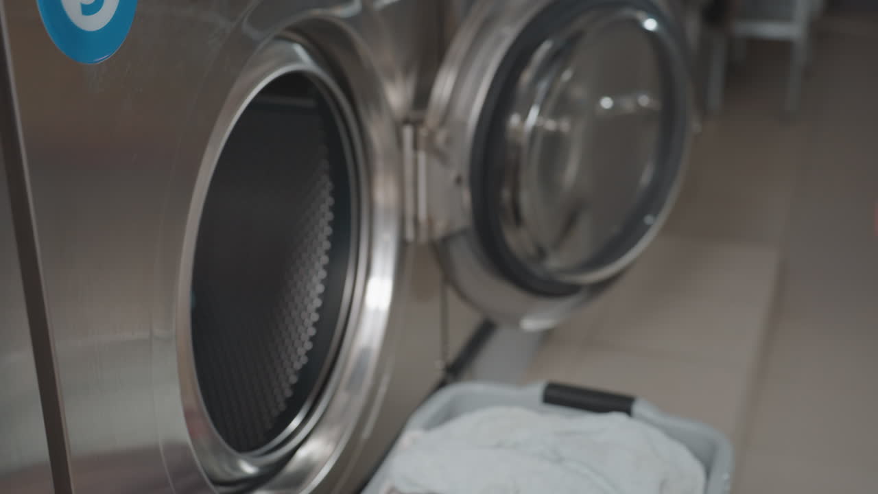 Aerial view of worker hand removing cloth from dryer, placing fabric into laundry basket, then moving across room, stainless drum with open door, hygienic service routine inside modern laundromat