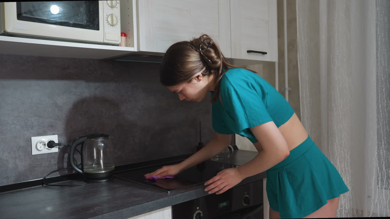 Donna caucasica appoggiata al piano di lavoro che pulisce la superficie con un panno, top corto e gonna color ottanio, faccende domestiche veloci, bollitore e forno a microonde sullo sfondo, scena domestica ordinata e pulita con azione mirata