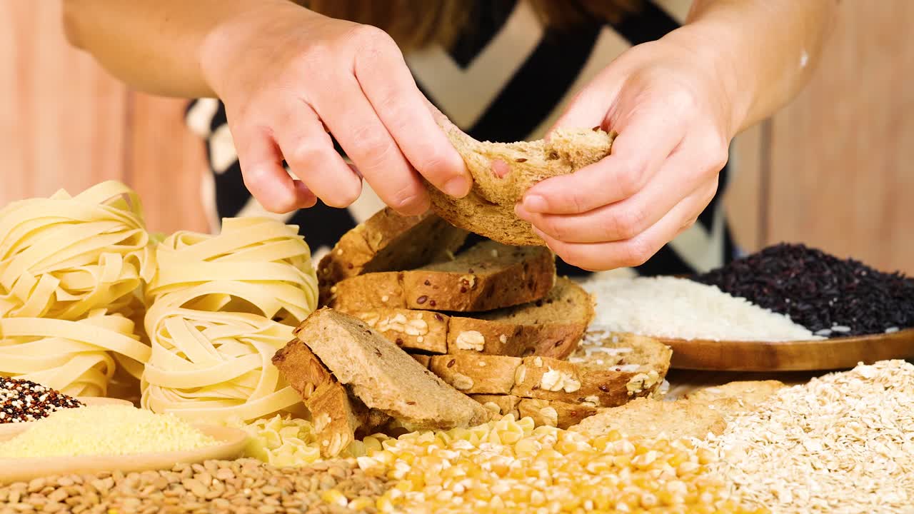 Hands tearing whole wheat bread surrounded by various grains and pasta. Bright lighting enhances the textures and colors