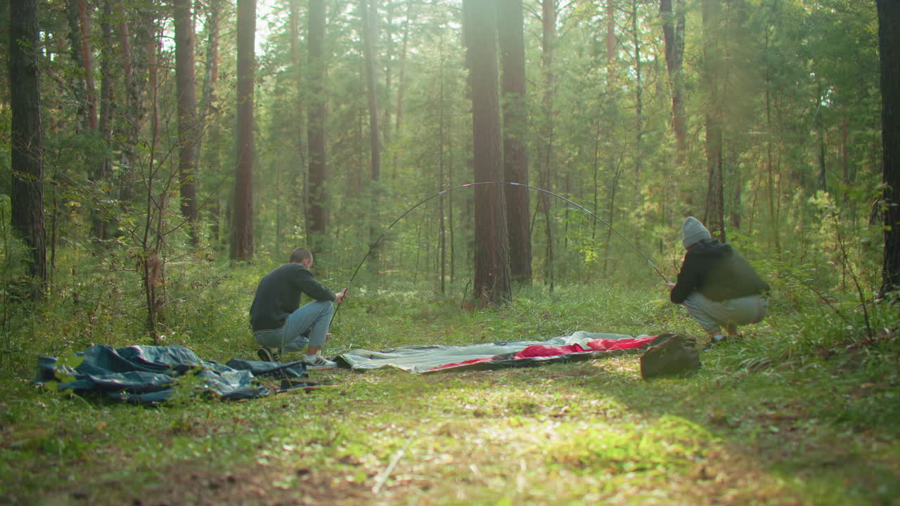 Couple prepares tent in forest clearing as man holds flexible tent pole and lady squats to adjust fabric, sunlight filters through trees creating natural glow over scene with tent bag visible nearby