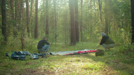 couple prepares tent in forest clearing as man holds flexible tent pole and lady squats to adjust fabric, sunlight filters through trees creating natural glow over scene with tent bag visible nearby