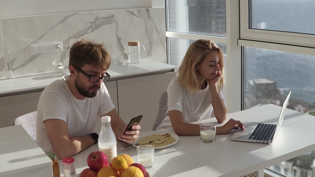 Couple's morning, they're sitting by a counter in kitchen, woman using laptop and man smartphone. Big panoramic windows with mild sunlight on background