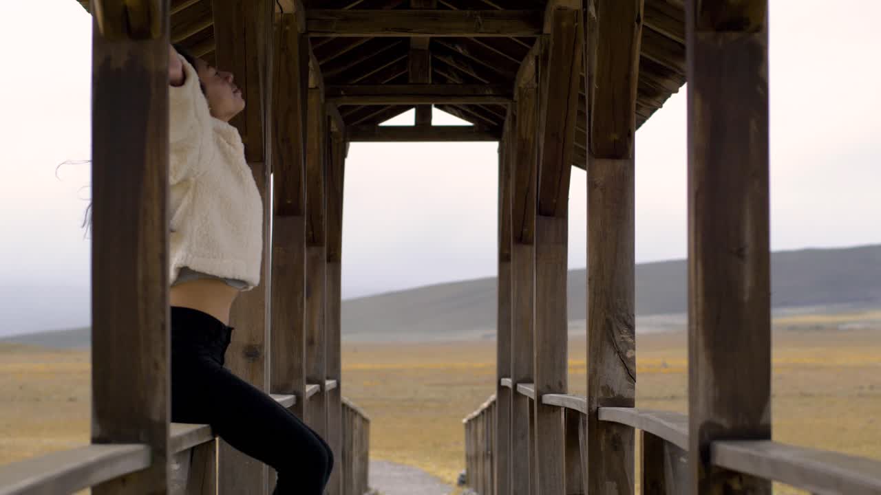 Dark hair tan woman sits on bridge railing lifting arms up and stretching as wind blows, ecuador