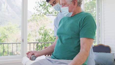 Senior-man-wearing-face-mask-with-male-therapist-while-exercising-with-dumbbells-at-retirement-home