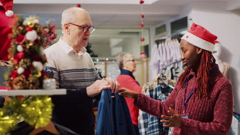 helpful retail assistant wearing santa hat during festive holiday season helping old man with best fitting blazer. employee assisting customer in christmas decorated clothing store