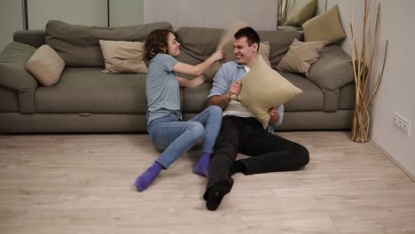 young caucasian couple having fun and enjoying quality time together sitting on the floor at home. couple is playing pillow fight close to the couch. slow motion