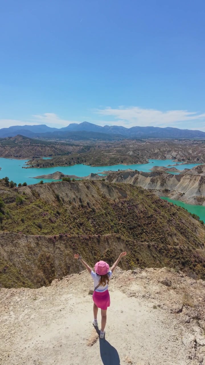 Vista aérea con dron de una viajera de pie en un mirador de acantilado con vistas a las aguas turquesas y las dramáticas tierras baldías del Embalse de Algeciras en Murcia, España