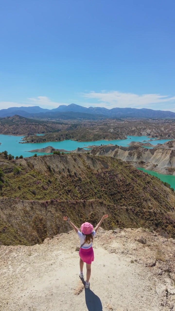 Vista aérea con dron de una viajera de pie en un mirador de acantilado con vistas a las aguas turquesas y las dramáticas tierras baldías del Embalse de Algeciras en Murcia, España