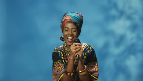 african american young woman in turban and traditional clothes singing with microphone and dancing while looking at camera