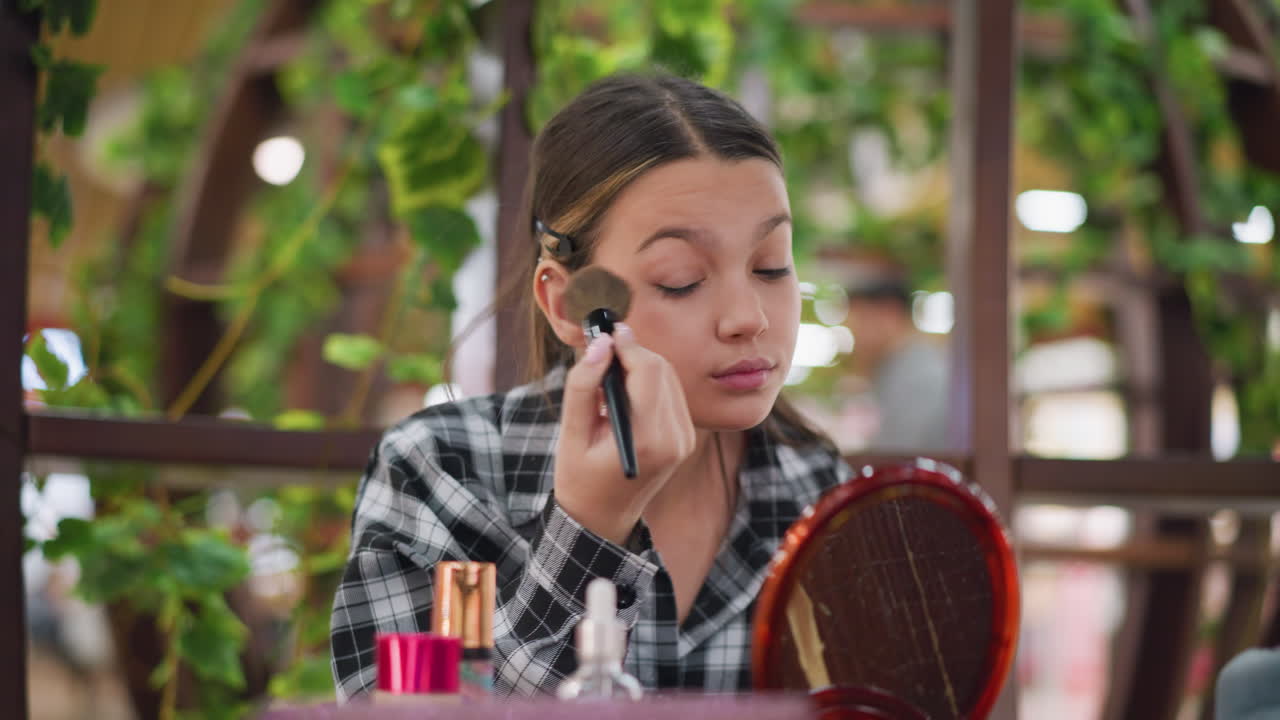 una joven se aplica maquillaje en el ojo usando un pincel de maquillaje mientras está sentada en la mesa, varios productos cosméticos como fundación, polvo y pincel de maquillaje se colocan en la mesa