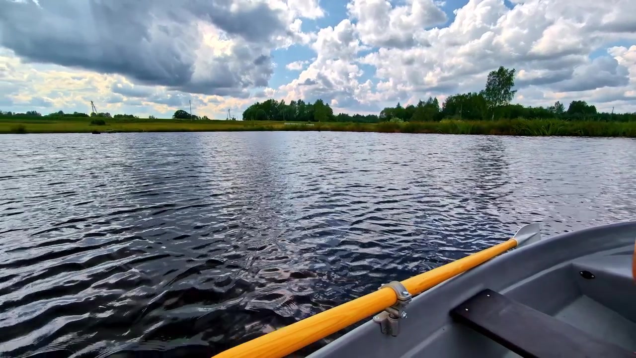 A rowboat gently floats on a calm river with rippling water and scattered clouds overhead near Cesis, Latvia, framed by green trees along the shore.