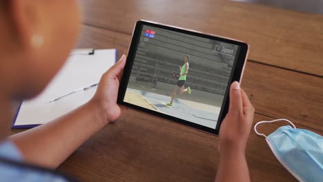 Composite-of-woman-sitting-at-table-with-face-mask,-watching-athletics-long-jump-event-on-tablet