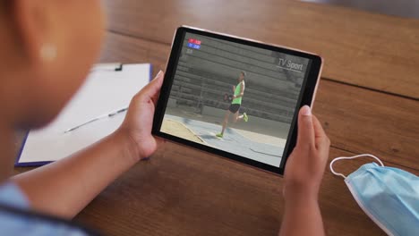 compuesto de una mujer sentada en una mesa con una máscara facial, viendo el evento de salto de longitud de atletismo en la tableta
