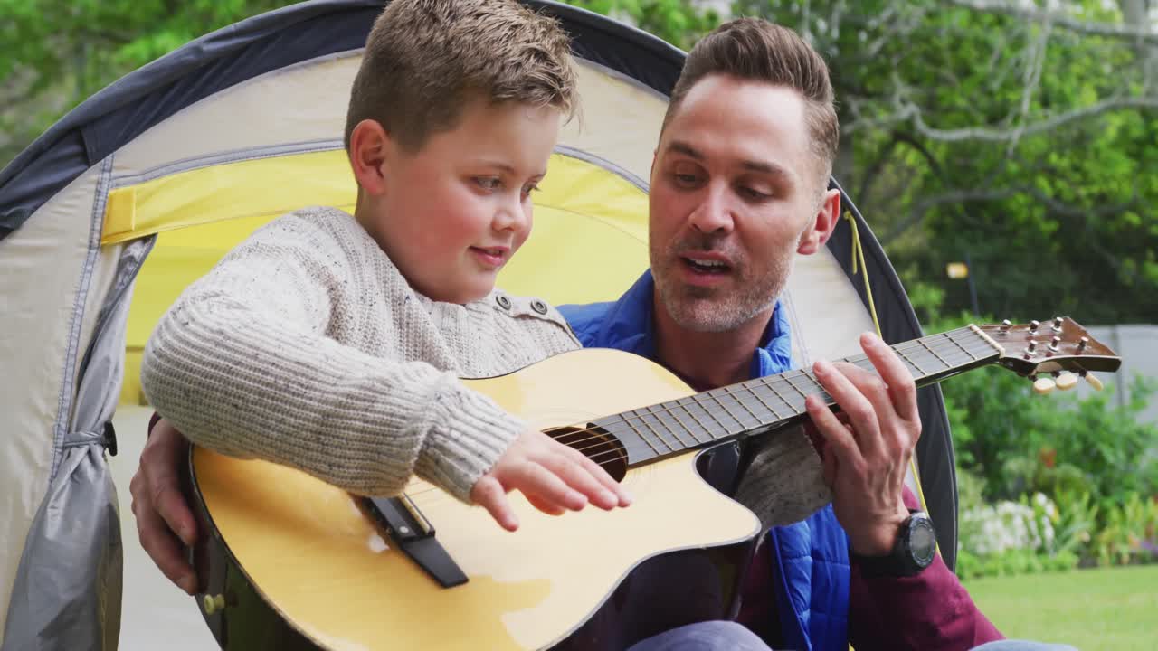 En glad kaukasisk far og søn, der sidder i et telt og spiller guitar i haven.