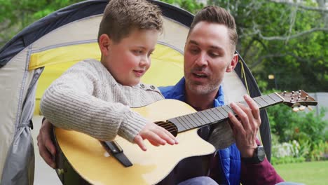 happy caucasian father with son sitting in tent and playing guitar in garden