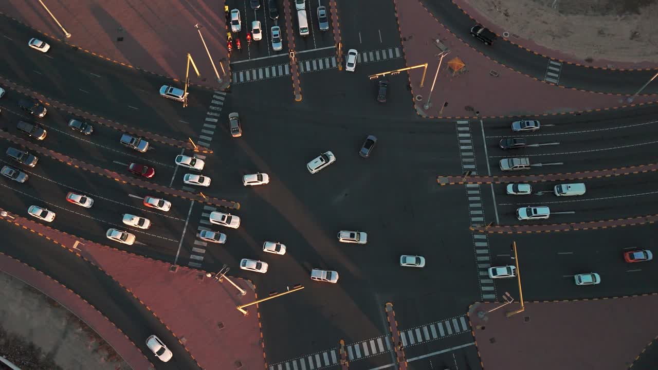 Top-down aerial of a busy multilane junction at golden hour; vehicles, zebra crossings and lane markings form a geometric urban pattern with long shadows across asphalt in a Gulf-region city