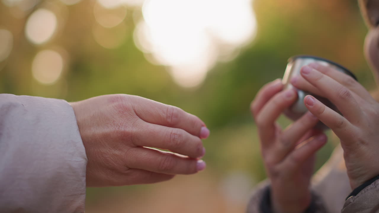 close up of adult hand offering cup of water to child reaching to drink, set against blurred autumn foliage background with warm bokeh and natural light, capturing caring gesture of refreshment