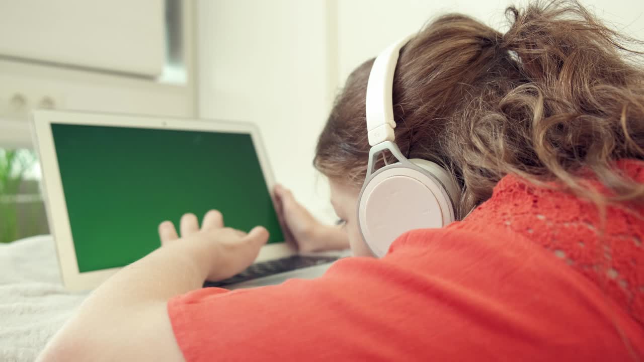 una chica cansada con auriculares se acuesta boca abajo en la cama y mira la pantalla de la computadora portátil.