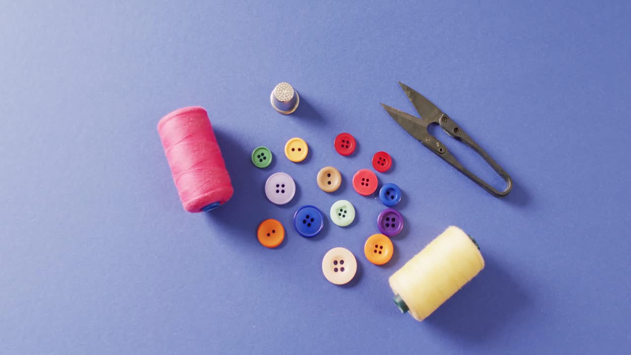 Video of thread snippers, thimble, buttons and reels of cotton on blue background