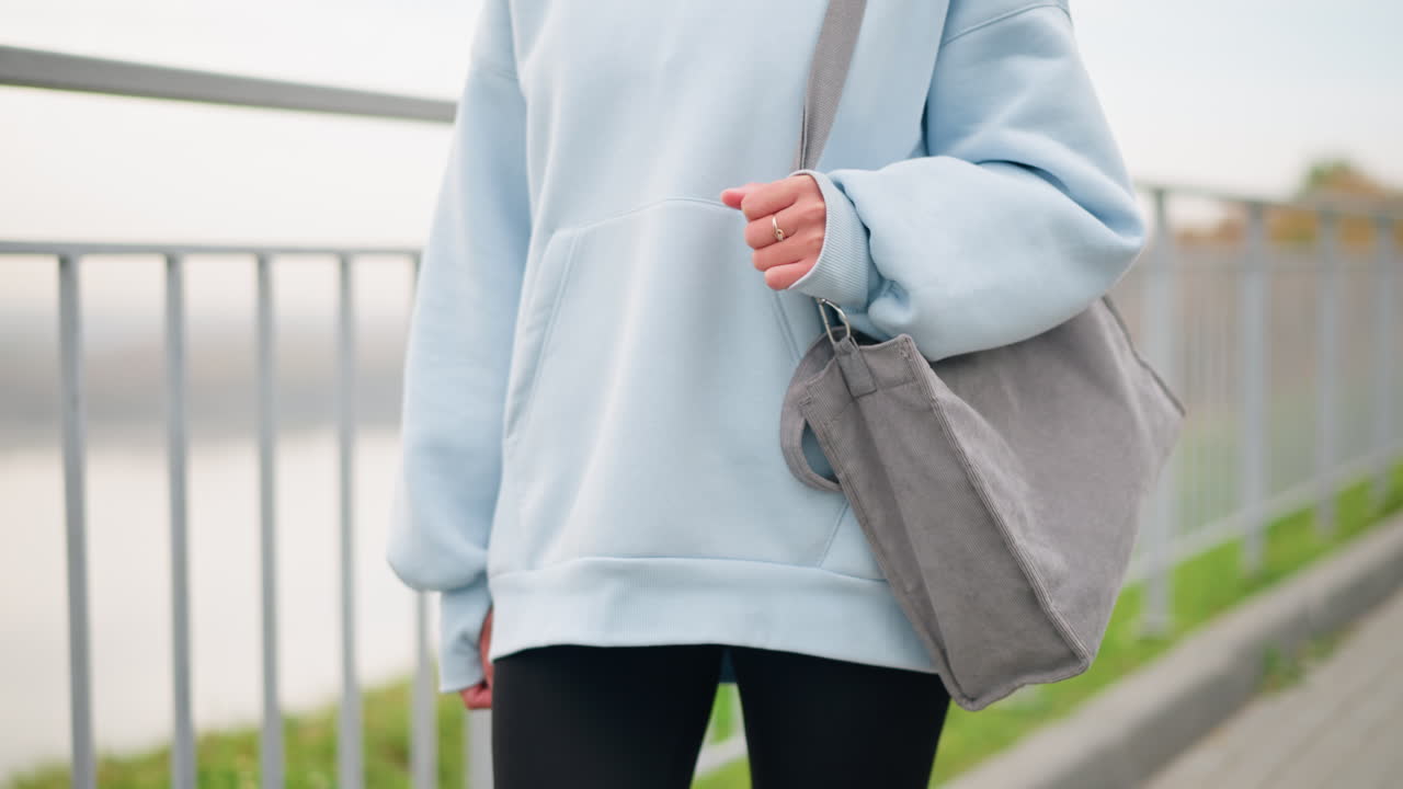 Partial view of young woman in casual blue hoodie walking with handbag and shiny golden ring on hand, with blurred iron fence and green grass in the background