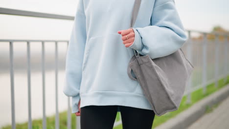 partial view of young woman in casual blue hoodie walking with handbag and shiny golden ring on hand, with blurred iron fence and green grass in the background