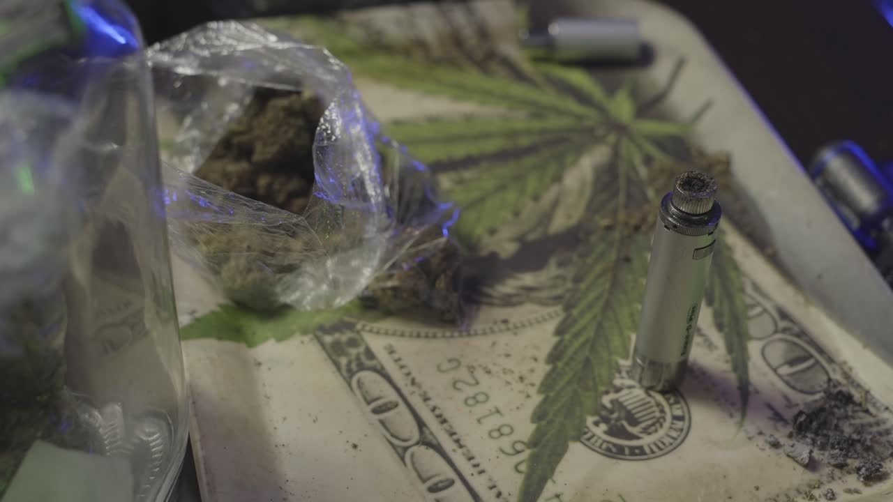 Close-up shot of a cannabis tray with buds, a half-used vape bowl, ash residue, and a dollar bill design with a leaf overlay.