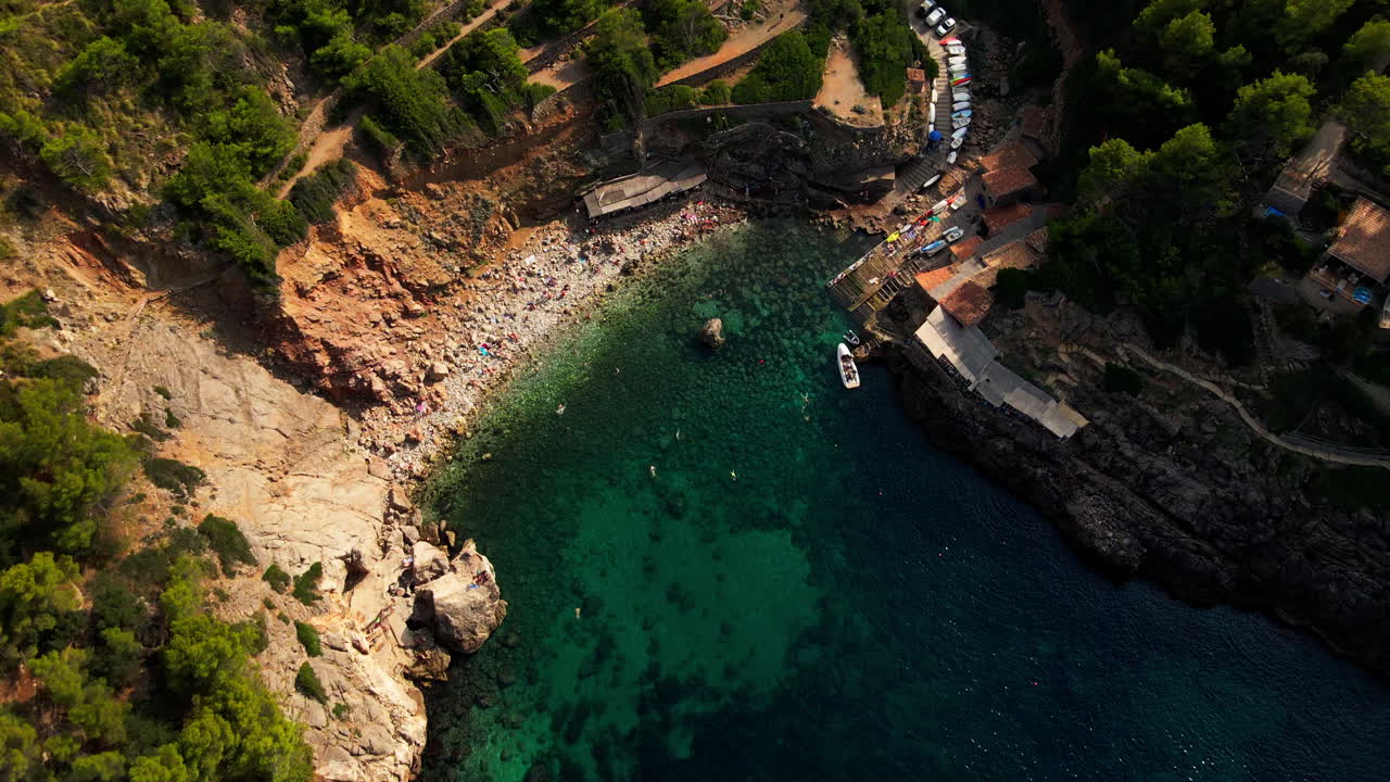 Spettacolare ripresa aerea in picchiata di una cala mediterranea isolata e una spiaggia con persone che nuotano e barche a remi.