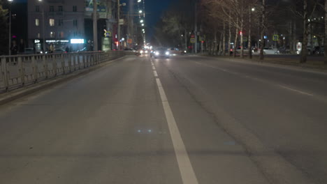 a nighttime city scene showing a wide, with people and car passing across the illuminated road by streetlights with car headlights approaching from the distance