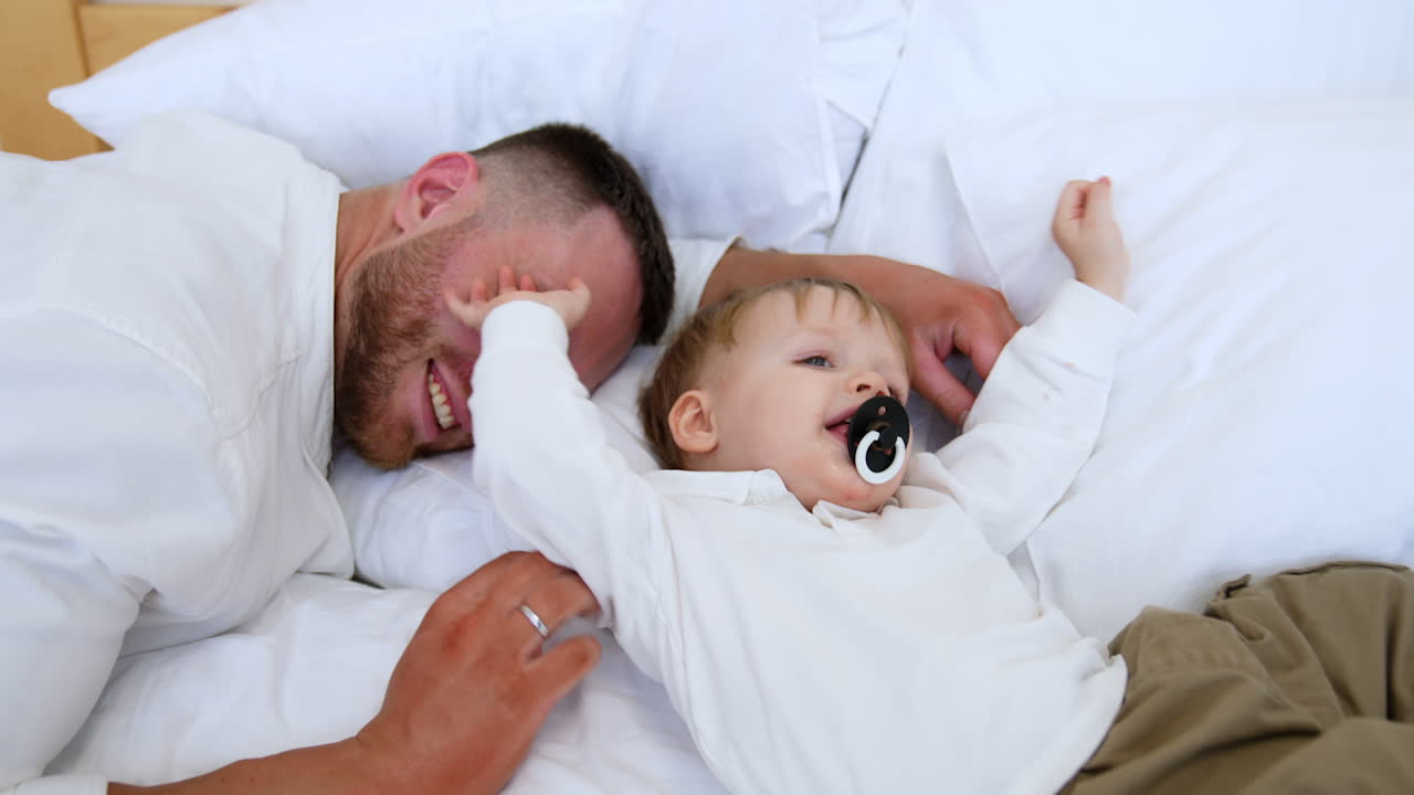 Cute little baby sucking a pacifier lies in the pillows. Bearded father lying beside smiles to kid. Toddler pushes his dad away.