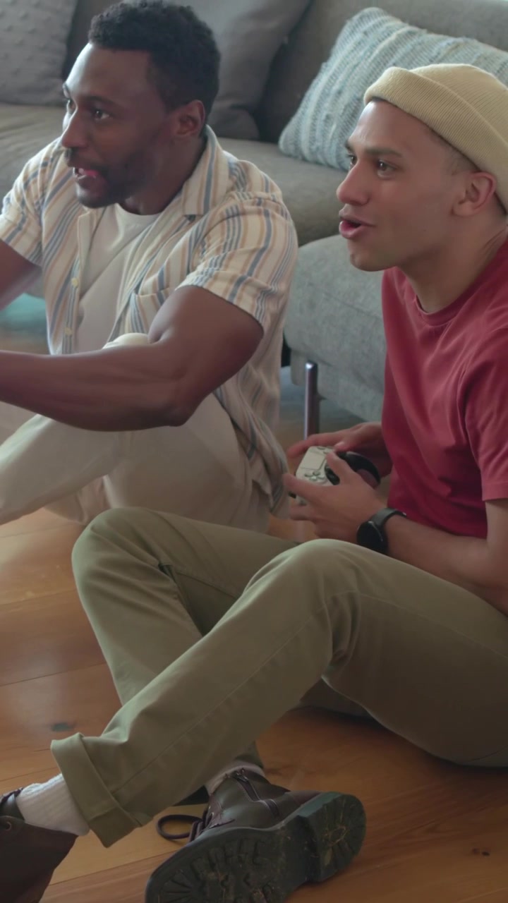 Vertical video: Leaning red tee man snacking and playing with friend at home with gamepads for fun