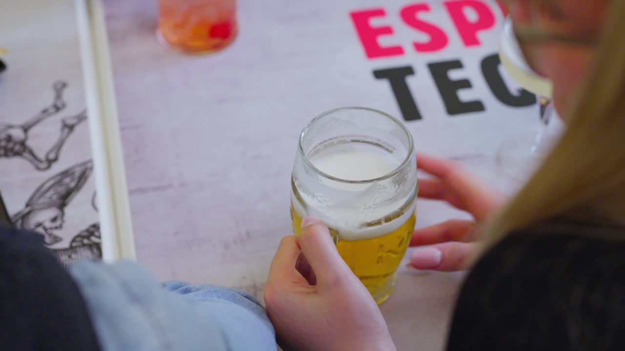 Hand on beer glass with frothy top at a bar table, casual atmosphere in the background