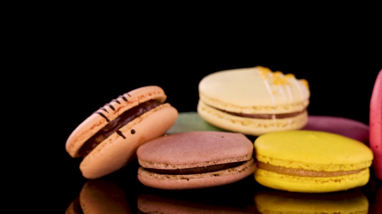 A vibrant selection of assorted macarons rotates on a glossy black surface, illuminated by studio lighting, highlighting their textures and pastel colors