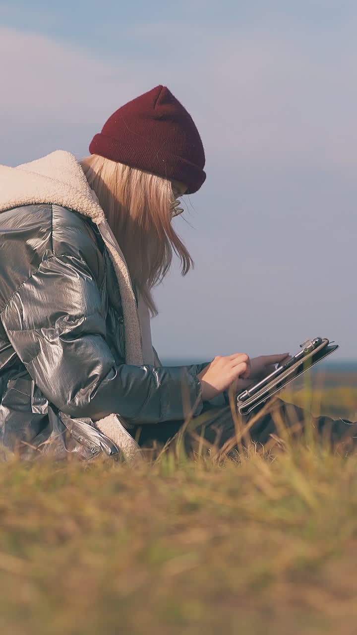 long haired girl in warn jacket works on tablet sitting by tent on autumn meadow against clear blue sky at calm sunset