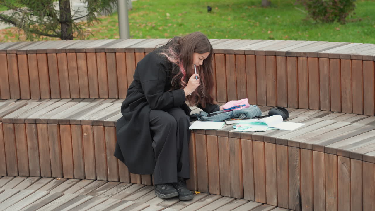 Ung kvinnelig student på trebenk som noterer og organiserer papirer ved siden av kamera og ryggsekk i høstparken, fokusert og ettertenksom atmosfære, lagvis kåpe, ensom studieøkt
