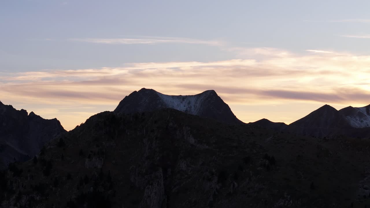 Ascesa aerea sopra una cresta retroilluminata con pareti rocciose in pendenza e un tenue bagliore giallo sulle nuvole contro il cielo blu.