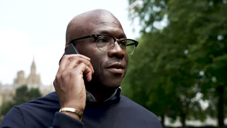 Bald-black-professional-male-stands-in-a-park-during-the-daytime,-talking-on-his-phone