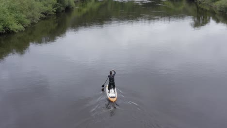 Aerial-follows-stand-up-paddleboarder-paddling-on-small-rural-river