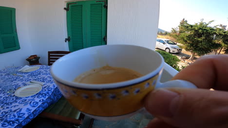 una persona camina con una taza de café en la mano afuera en una terraza antes de sentarse a la mesa