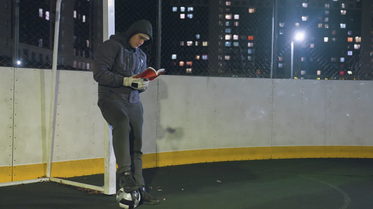 하밤에 금속 골포스트에 기대어 축구공을 고 야외에서 책을 읽는 남자, 도시 불빛으로 둘러싸여