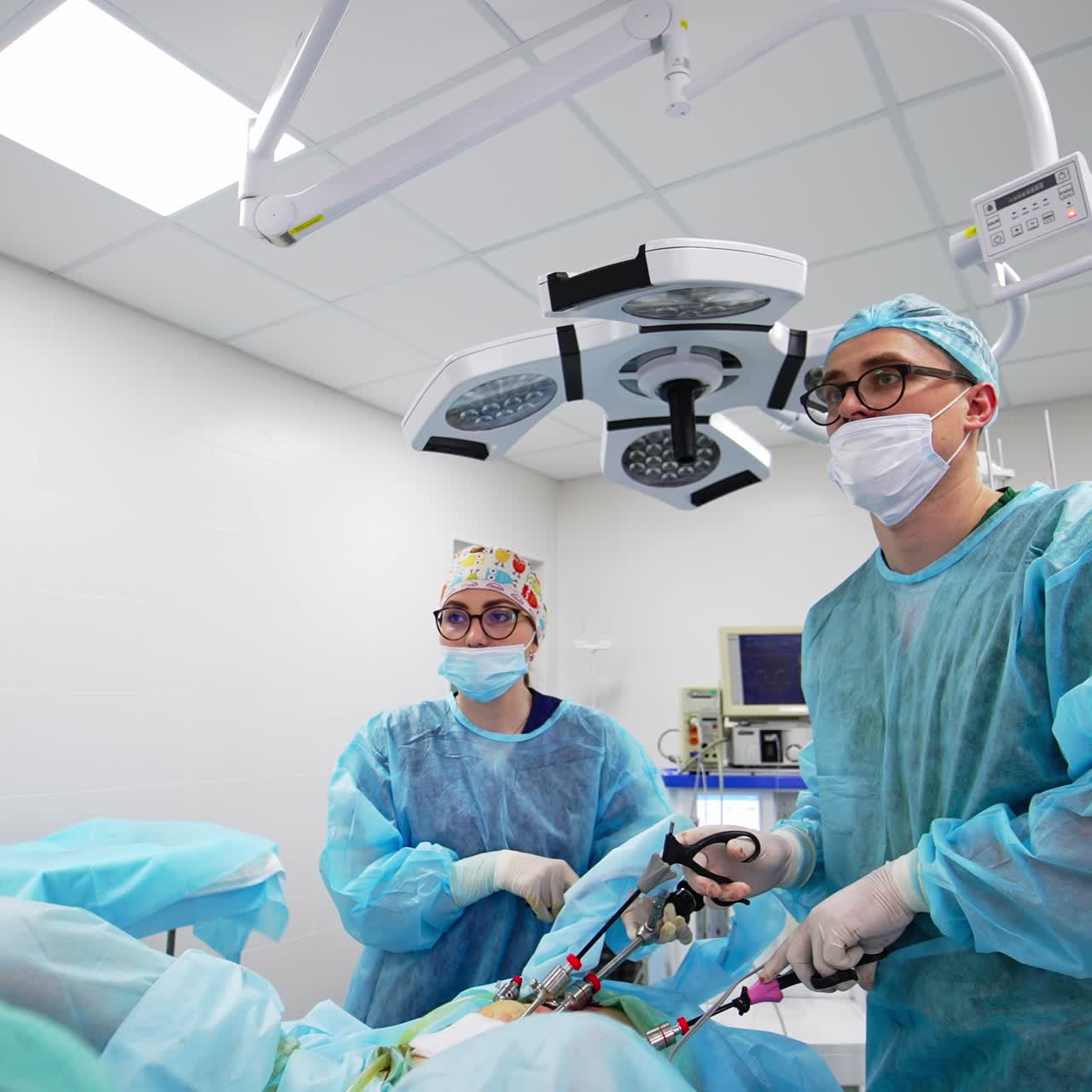 Young male doctor applying instruments at operation. Female assistant standing beside. Modern surgery room backdrop