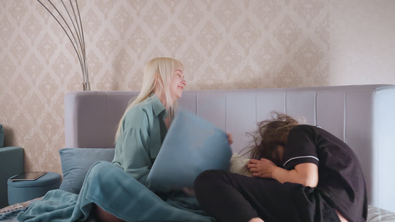 Two girls enjoy playful pillow fight while sitting on bed in bright bedroom, one girl smiles while swinging blue pillow, other girl defends with white one