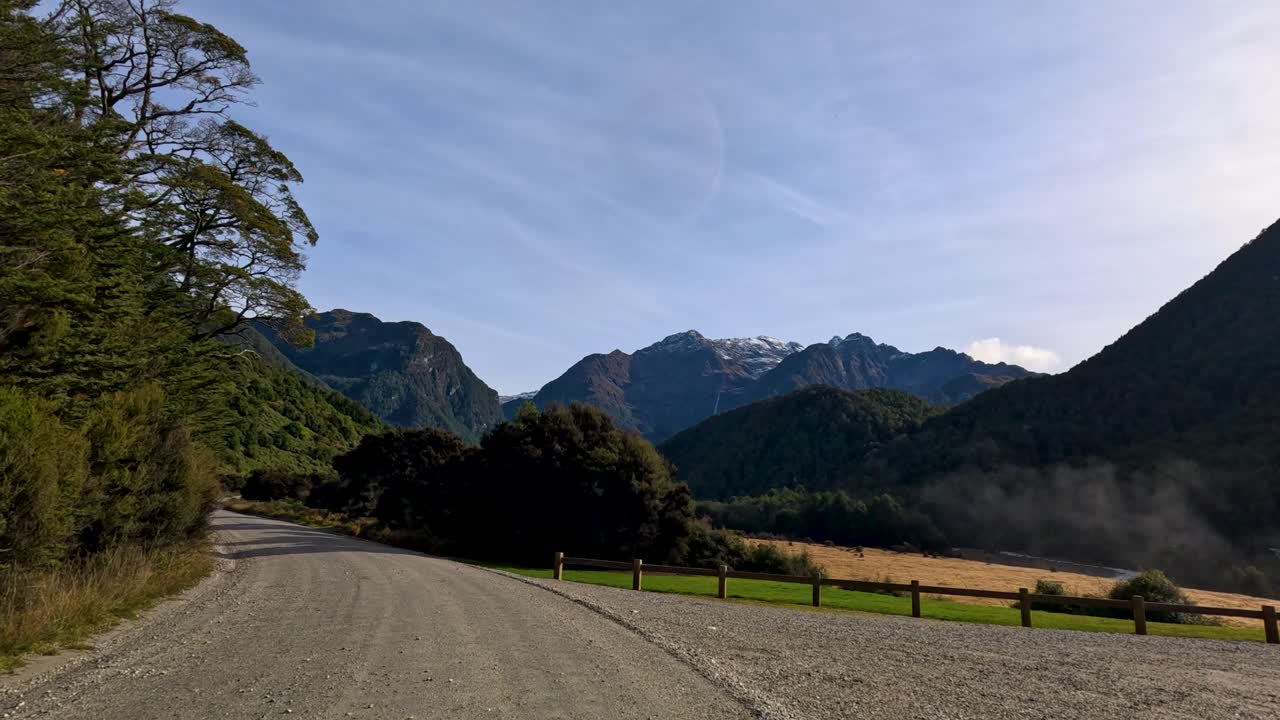 Vehicle travels gravel road through lush forest, mountain scenery, and open landscape in daylight