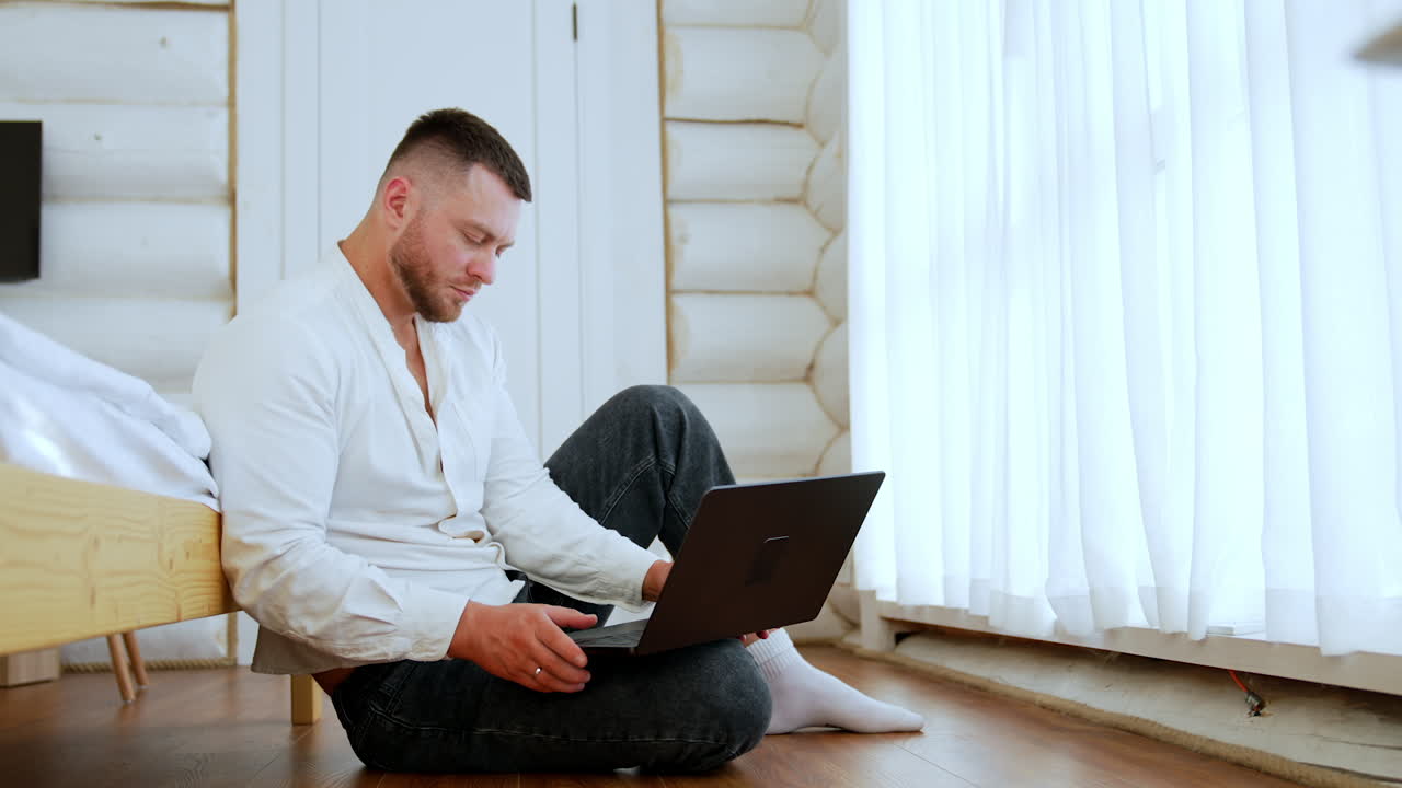 Focused serious Caucasian man sits on the floor holding a laptop. Man surfs the internet or works online.