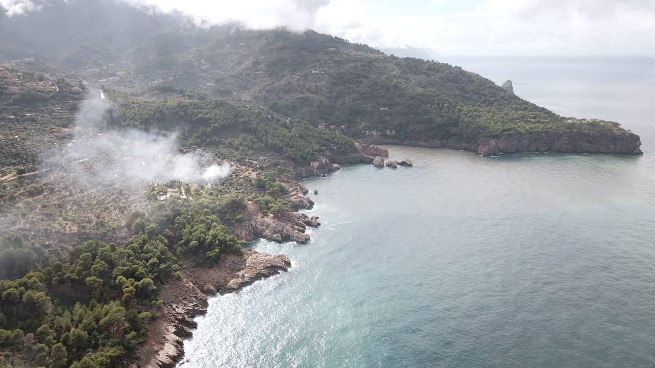 La telecamera aerea si muove in avanti lungo le scogliere ricoperte di pini, oltrepassando un villaggio collinare con fumo che si disperde e rivelando un'insenatura riparata sulla costa di Tramuntana a Maiorca