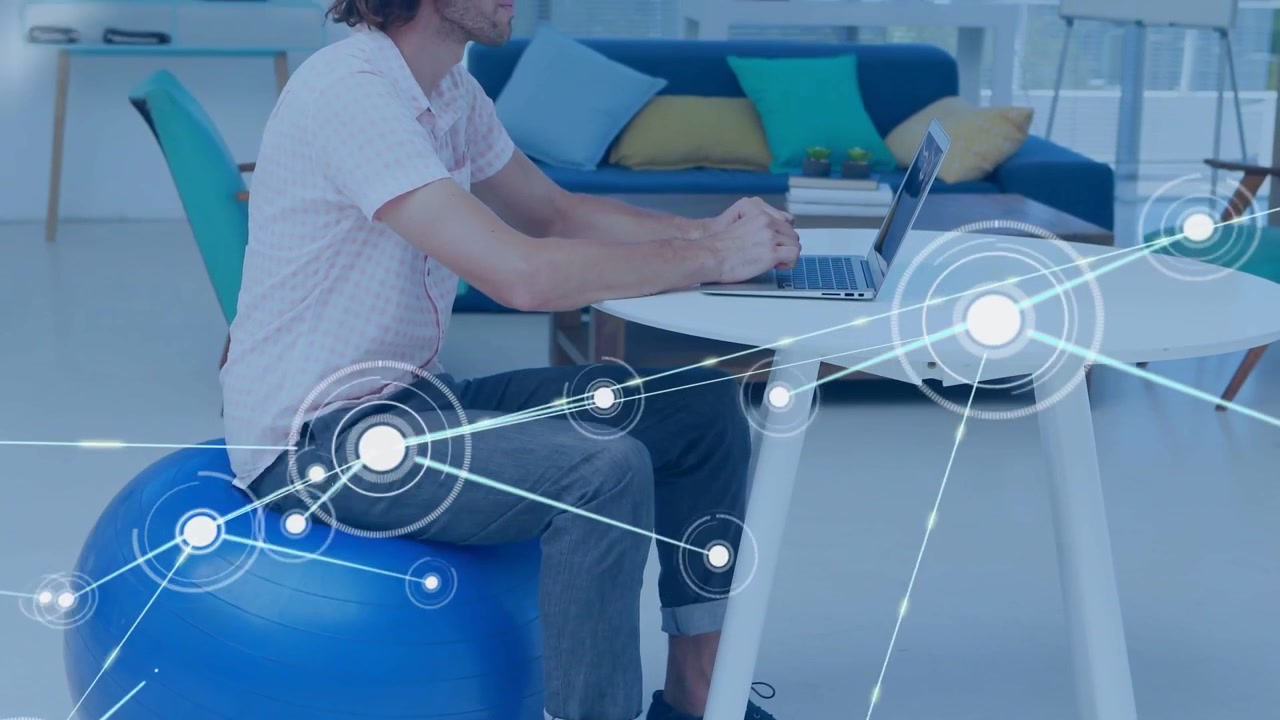 Typing man in short-sleeve shirt in modern lounge with blue exercise ball, laptop and glowing nodes