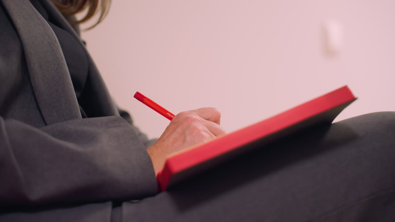 Nærbilde av forretningskvinne som skriver i notatbok med rød penn, sittende på stol i formell antrekk, fokusert på arbeid eller notater, kreativt og profesjonelt miljø, forretningskonsept
