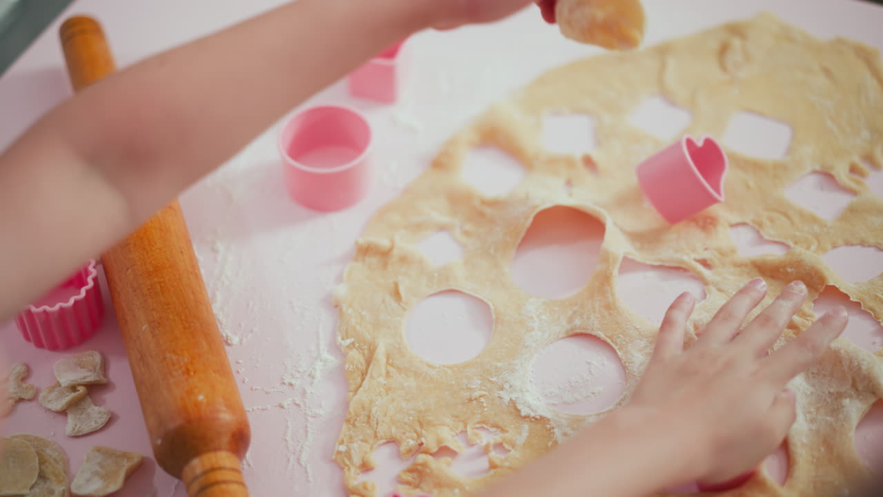 Boy removes circular shape from flattened dough using pink cutter while another child works in background, hands dusted with flour on pastel pink table creating playful baking scene
