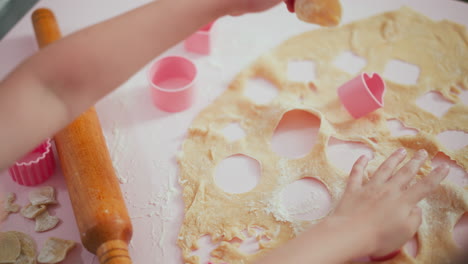 boy removes circular shape from flattened dough using pink cutter while another child works in background, hands dusted with flour on pastel pink table creating playful baking scene
