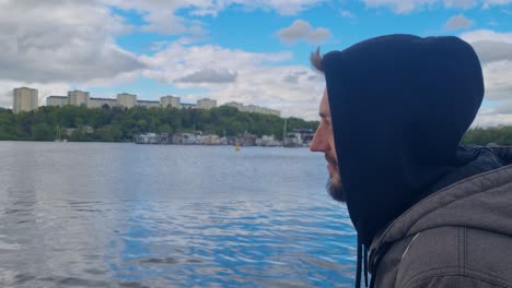 A man in a hoodie gazes at the water in Stockholm on a cloudy spring evening, calmly meditating, contemplating life, and enjoying his existence in the crisp Nordic air