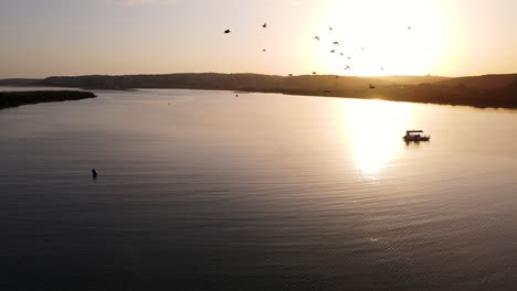 Antena-Cinematográfica-De-Puesta-De-Sol-De-La-Silueta-Del-Barco-En-Reflejo-Dorado-Y-Amarillo-En-La-Superficie-Del-Estuario-Del-Río-Keurbooms,-Aves-Acuáticas-Que-Pasan-Volando